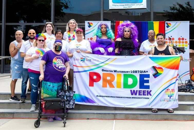 Group of diverse LGBTQIA+ individuals posing in front of a banner that reads 'Greater Waterbury LGBTQIA+ Pride Week', with colorful decorations in the background.